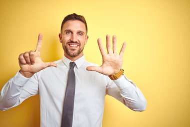 Sarı izole edilmiş arka plan üzerinde zarif beyaz gömlek giyen genç yakışıklı iş adamı yedinci parmaklarıyla işaret ederken kendinden emin ve mutlu bir şekilde gülümsüyor..