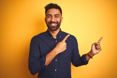 Genç ve yakışıklı Hintli işadamı, izole edilmiş sarı arka plan üzerinde gömlek giyiyor gülümsüyor ve kameraya bakıyor. İki eliyle ve parmaklarıyla kenarı işaret ediyor..