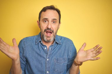 Sarı arka plan üzerinde gri saçlı, yakışıklı, orta yaşlı bir adam. Kolları ve elleri havada, kafası karışmış. Şüphe kavramı.