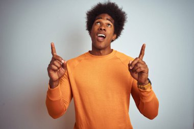 Afro saçlı, turuncu süveter giyen genç bir Amerikalı beyaz arka planda şaşkın şaşkın yukarı bakıyor ve parmaklarıyla işaret ediyor ve kollarını kaldırıyordu..