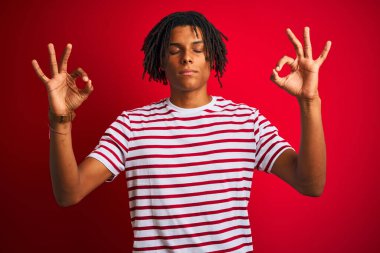 Rastalı tişörtlü genç afro adam izole edilmiş kırmızı arka planda duruyor rahatlamış ve gözleri kapalı meditasyon hareketi yaparken parmaklarıyla gülümsüyor. Yoga konsepti.