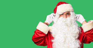 Orta yaşlı yakışıklı adam, Noel Baba kostümü ve sakal takmış gülümsüyor parmaklarıyla kulaklarını çekiyor, komik bir jest. Seçme sorunu