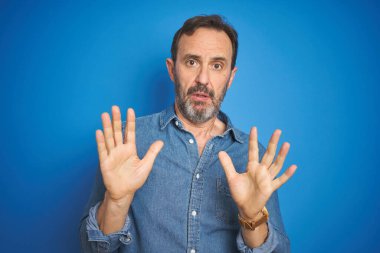 İzole edilmiş mavi arka plan üzerinde gri saçlı yakışıklı orta yaşlı bir adam. Avuçlarını kaldırıyor, reddediyor ve iğrenç bir ifadeyle reddediyor. Dur ve yasakla.