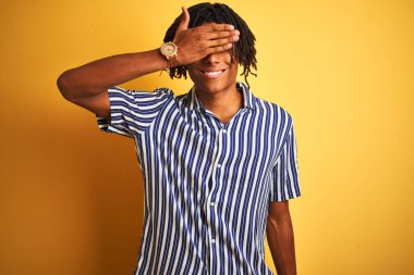 Rastgele çizgili tişörtü olan Afro bir adam izole edilmiş sarı arka planda gülümseyen ve yüzünde şaşkınlık için gözleri kapayarak gülen bir adam. Kör kavram.