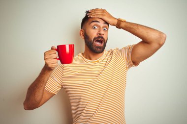 İzole edilmiş beyaz arka planda duran kırmızı kahve içen genç Hintli adam, elleri başının üstünde, utanç ve şaşkınlık dolu, sinirli ve hayal kırıklığına uğramış bir halde. Hata için korku ve üzüntü.