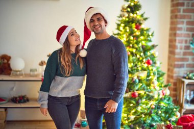 Mutlu ve kendinden emin gülümseyen genç bir çift. Evde Noel Baba şapkası takmış Noel ağacının etrafında dikiliyor.