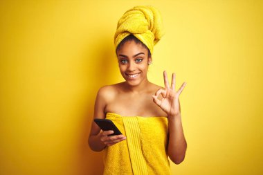Duş üstüne havlu giyen Afro kadın izole edilmiş sarı arka plan üzerinde smatrphone kullanıyor. Tamam işareti yapıyor, mükemmel bir sembol.