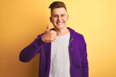 Mor kazak giyen genç yakışıklı adam izole edilmiş sarı arka planda duruyor mutlu bir el hareketi yapıyor. Kameraya bakıp başarıyı gösteren ifade onaylanıyor.