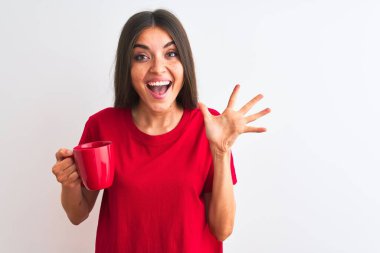Genç güzel kadın izole beyaz arka plan üzerinde duran kahve kırmızı fincan içme çok mutlu ve heyecanlı, kazanan ifade büyük bir gülümseme ile çığlık zafer kutluyor ve ellerini kaldırdı