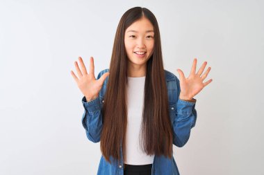 İzole edilmiş beyaz arka planda duran kot gömlekli genç Çinli kadın on numarayı gösterirken kendinden emin ve mutlu bir şekilde gülümsüyor..