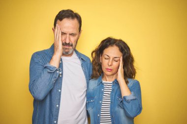 Güzel bir orta yaşlı çift, izole edilmiş sarı arka planda kot gömlek giyiyorlar ve stres yüzünden başları ağrıyordu. Migren ağrısı..