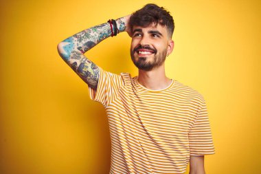 İzole edilmiş sarı arka plan üzerinde duran çizgili tişörtlü genç adam. Kendine güvenen, el hareketleriyle saçlarına dokunan, çekici ve modaya uygun poz veren.