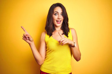 Sarı izole arka plan üzerinde ayakta t-shirt giyen genç güzel kadın gülümseyen ve yan iki el ve parmak ile işaret kamera bakıyor.