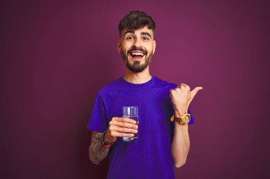 Dövmeli genç adam izole edilmiş mor arka planda duran su bardağını içerken başparmağıyla gülümseyerek yüzünü gösteriyor.