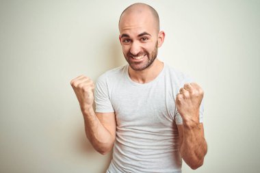 Kel ve sakallı genç bir adam izole edilmiş arka planda günlük beyaz tişört giyiyor çok mutlu ve heyecanlı bir şekilde kollarını kaldırıp, gülümseyerek ve başarı için çığlık atarak zafer hareketi yapıyor. Kutlama konsepti.