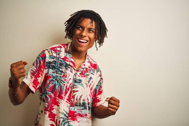 Tatilde rastalı bir Afro adam izole edilmiş beyaz arka plan üzerine yazlık gömlek giyiyor çok mutlu ve heyecanlı bir şekilde kollarını kaldırıp, gülümseyerek ve başarı için çığlık atarak kazanan jestler yapıyor. Kutlama konsepti.