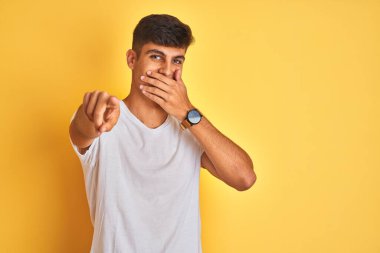 İzole edilmiş sarı arka planda duran beyaz tişörtlü genç Hintli adam sana gülüyor, kamerayı işaret ediyor, ağzının üzerinde utanç ifadesi var.