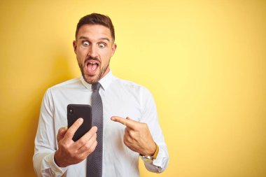 Genç ve yakışıklı bir iş adamı sarı arka plan üzerinde akıllı telefon kullanıyor. Elle ve parmakla işaret ederek çok mutlu.