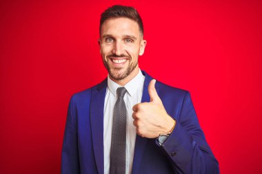 Genç ve yakışıklı bir iş adamı kırmızı arka planda mutlu bir el hareketi yapıyor. Kameraya bakıp başarıyı gösteren ifade onaylanıyor.