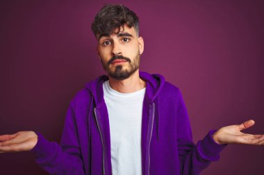 Çizgili polo giyen dövmegiyen genç adam izole sarı arka plan üzerinde duran habersiz ve kolları ve elleri ile karışık ifade kaldırdı. Şüphe kavramı.