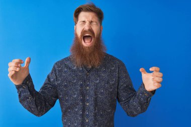 Çiçekli yaz gömleği giyen genç kızıl saçlı adam izole edilmiş mavi arka planda duruyor ve başarı için çıldırıyor kollarını kaldırmış ve gözlerini kapatmış heyecanla haykırıyor. Kazanan konsept