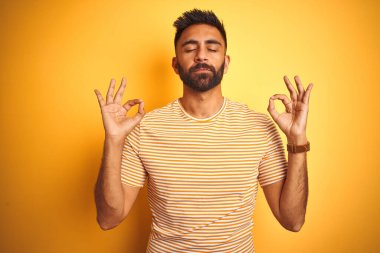 İzole edilmiş sarı arka plan üzerinde duran genç Hintli adam rahatlamış ve gözleri kapalı meditasyon hareketi yaparken parmaklarıyla gülümsüyor. Yoga konsepti.