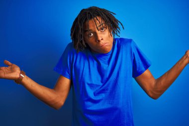 Rastalı tişörtlü Afro-Amerikan bir adam izole edilmiş mavi arka plan üzerinde duruyor. Kolları ve elleri havada, kafası karışmış bir ifade. Şüphe kavramı.