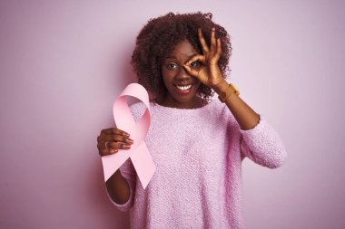 Kanserli kurdele tutan Afrikalı genç bir kadın mutlu yüzüyle pembe arka planda duruyor. El ele tutuşup parmaklarının arasından bakıyor.