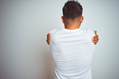İzole edilmiş beyaz arka planda tişört giyen genç Hintli adam kendine mutlu ve pozitif bir şekilde sarılıyor. Sevgi ve özgüven.