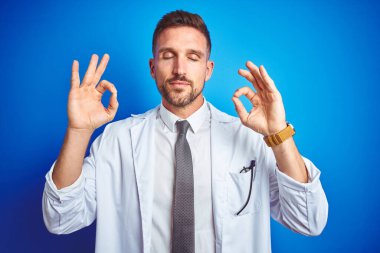 İzole edilmiş arka plan üzerinde beyaz profesyonel ceket giyen genç yakışıklı doktor rahatla ve parmaklarıyla meditasyon hareketi yaparken gözleri kapalı gülümse. Yoga konsepti.