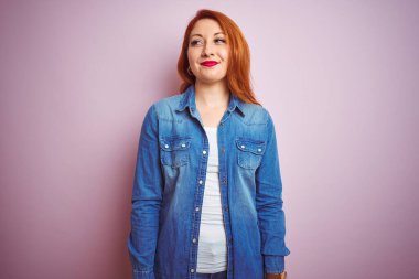 Genç, güzel, kızıl saçlı, kot gömlek giyen, izole edilmiş pembe arka planda duran, gülümseyen ve başka tarafa bakıp düşünen bir kadın..
