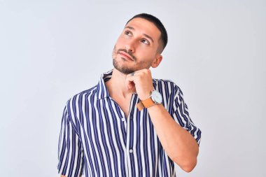 Deniz çizgili gömlek giyen genç yakışıklı bir adam izole arka planda duruyor, elleri çenesinde soru sorarken, düşünceli bir ifade takınıyor. Düşünceli bir yüzle gülümsüyor. Şüphe kavramı.