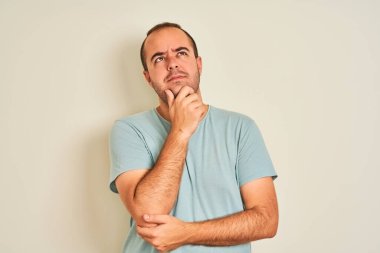 Mavi günlük tişört giyen genç bir adam, izole edilmiş beyaz arka planda duruyor, elleri çenesinde soru sorarken, düşünceli bir ifade takınıyor. Düşünceli bir yüzle gülümsüyor. Şüphe kavramı.