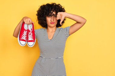Kıvırcık saçlı genç Arap kadın izole edilmiş sarı arka plan üzerinde spor ayakkabıları tutuyor. Kızgın yüz ifadesi, olumsuz işaret baş parmakları aşağıda, reddedilme kavramı.