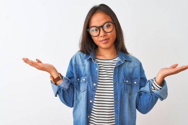 İzole edilmiş beyaz arka plan üzerinde kot gömlek ve gözlük takan Çinli genç bir kadın. Kolları ve elleri havada, kafası karışmış bir ifade. Şüphe kavramı.