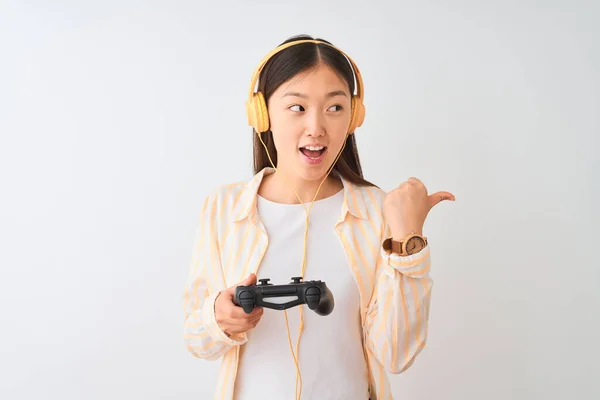 中国游戏女玩家用耳机在孤立的白色背景下玩电子游戏 手指朝天 笑容满面 — 图库照片