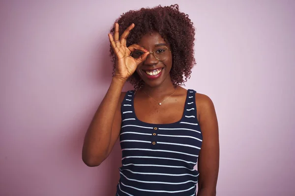 Young african afro woman wearing striped t-shirt glasses over isolated pink background doing ok gesture with hand smiling, eye looking through fingers with happy face.