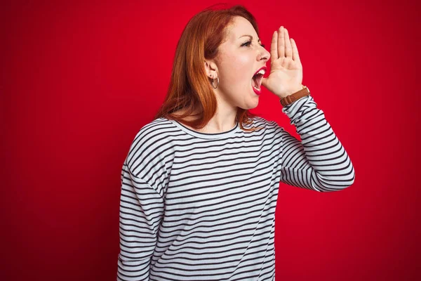 Young redhead woman wearing strapes navy shirt standing over red isolated background shouting and screaming loud to side with hand on mouth. Communication concept.