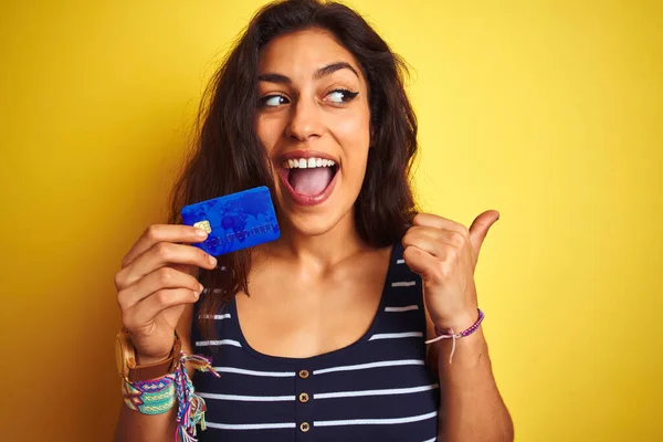 Young beautiful woman holding credit card standing over isolated yellow background pointing and showing with thumb up to the side with happy face smiling