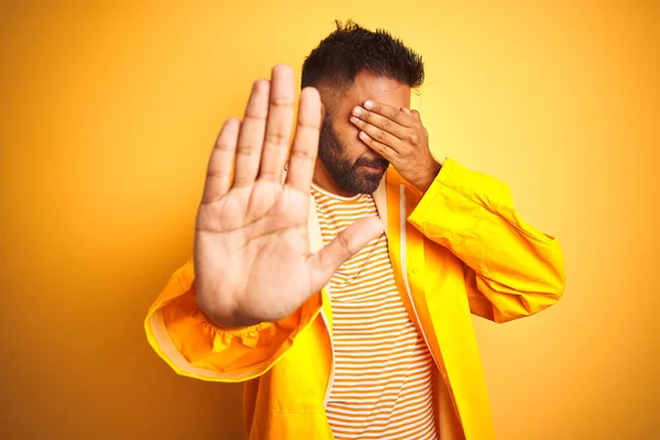 Young indian man wearing raincoat standing over isolated yellow background covering eyes with hands and doing stop gesture with sad and fear expression. Embarrassed and negative concept.