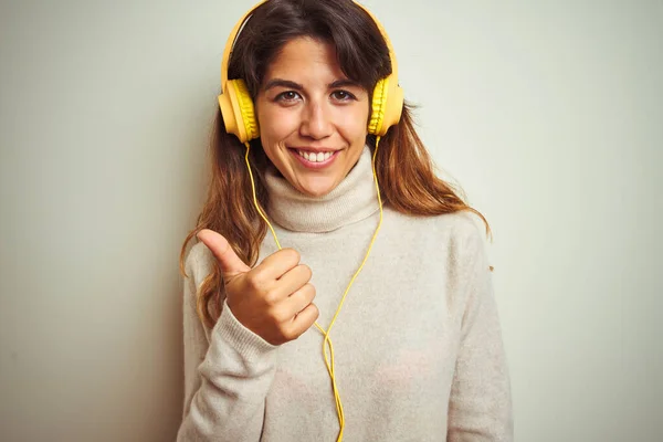 年轻美丽的女人在白色的背景下 用耳机听着音乐 手举着快乐的大拇指做手势 用成功的表情看着摄像机 — 图库照片