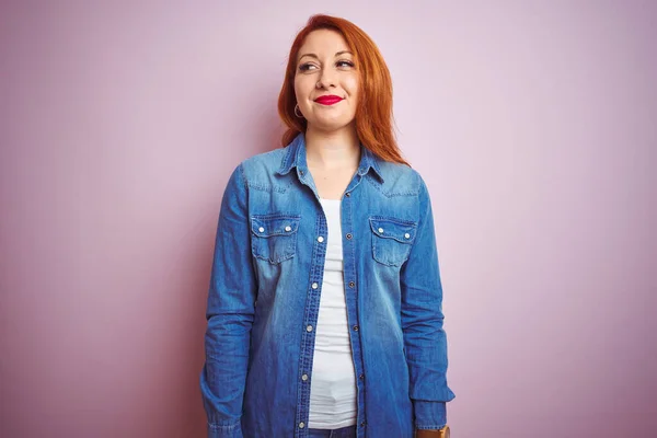 Youg beautiful redhead woman wearing denim shirt standing over isolated pink background smiling looking to the side and staring away thinking.