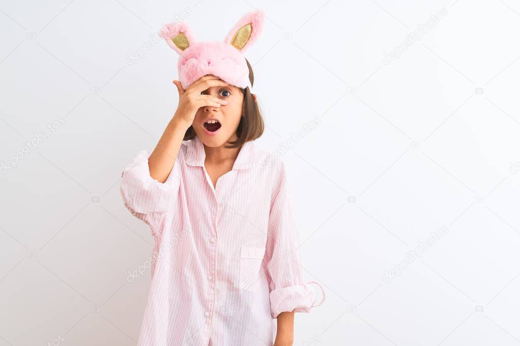 Beautiful child girl wearing sleep mask and pajama standing over isolated white background peeking in shock covering face and eyes with hand, looking through fingers with embarrassed expression.