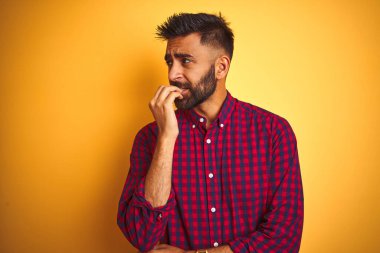 İzole edilmiş sarı arka planda dikilen rahat gömlekli genç Hintli adam elleri ağız yiyen tırnaklarla gergin ve stresli görünüyor. Endişe sorunu.