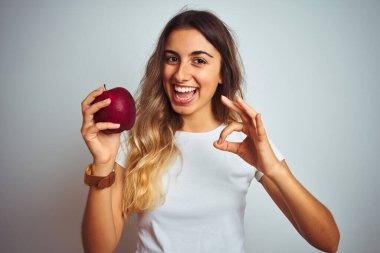 Gri izole arka planda kırmızı elma yiyen güzel bir kadın. Tamam işareti yapıyor. Parmaklarıyla, mükemmel bir sembol.