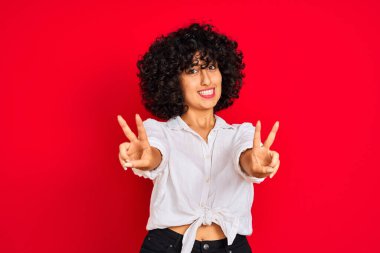 Kıvırcık saçlı, beyaz gömlekli, izole edilmiş kırmızı arka planda gülümseyen genç Arap kadın parmaklarını zafer işareti yaparken kameraya bakıyor. İki numara..