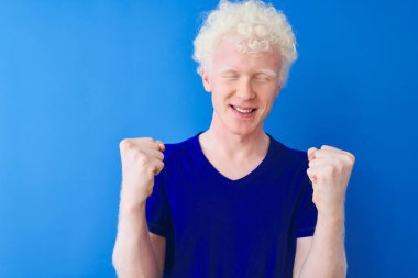 Sıradan tişörtler giyen genç albino bir adam izole edilmiş mavi arka planda durup kollarını kaldırarak ve açık gözlerle başarının şaşkınlığını ve şaşkınlığını kutluyor. Kazanan konsept.