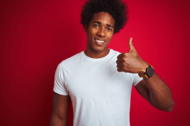 Afro saçlı, beyaz tişörtlü, izole edilmiş kırmızı arka planda duran genç bir Amerikalı. Elle mutlu bir jest yapıyor. Kameraya bakıp başarıyı gösteren ifade onaylanıyor.