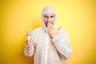 Boyama ekipmanı giyen ve elinde boya fırçası olan genç bir adam izole edilmiş sarı arka plan kaplamalı ağız hata yüzünden utançla dolu, korku ifadesi, sessizlikten korkmak, gizli konsept.