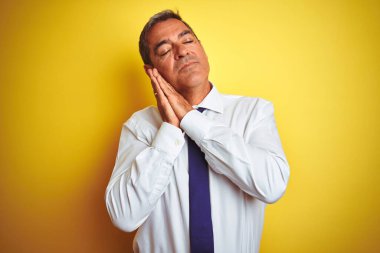 Yakışıklı orta yaşlı işadamı, izole edilmiş sarı arka planda duruyor. Yorgun uyuyor, hayal kuruyor ve ellerimiz kapalı gülümserken poz veriyor..